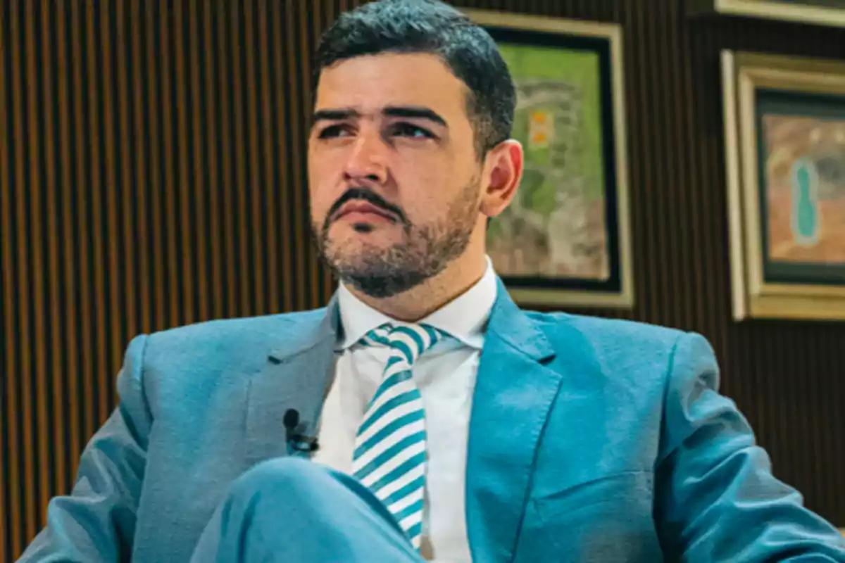 Hombre con barba y cabello corto vestido con traje azul claro y corbata de rayas sentado en una oficina con cuadros en la pared