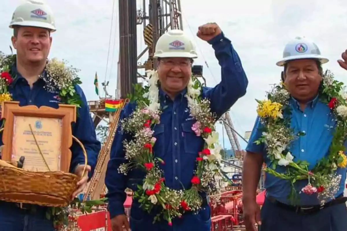 Three men wearing safety helmets and blue jerseys smile as they pose with flower garlands around their necks in an outdoor industrial setting; one of them holds a framed diploma and a basket, while another raises his fist in celebration. Three men wearing safety helmets and blue jerseys smile as they pose with flower garlands around their necks in an outdoor industrial setting; one of them holds a framed diploma and a basket, while another raises his fist in celebration.