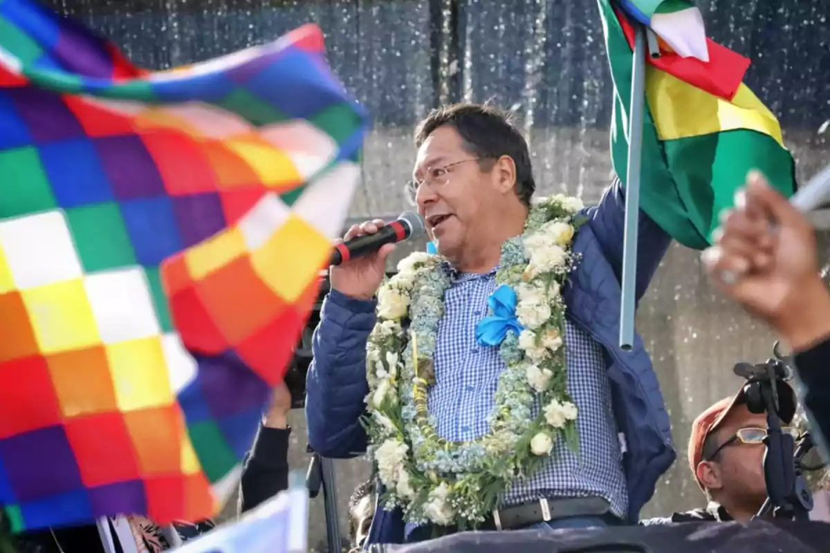 Hombre con guirnalda de flores hablando por micrófono y sosteniendo una bandera boliviana en un evento público rodeado de banderas multicolores