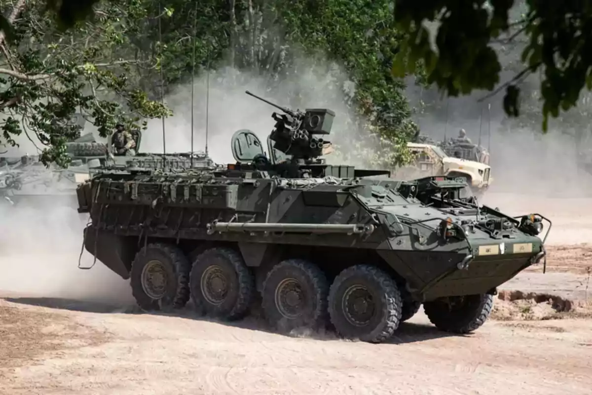 Eight-wheeled armored military vehicle advancing along a dirt road surrounded by trees and dust