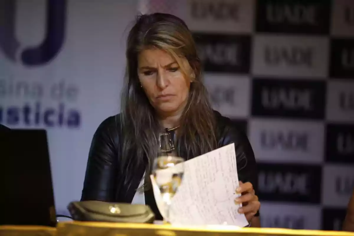 Mujer de cabello largo y rubio con expresión seria revisando unas hojas manuscritas en una mesa, con fondo de logotipos de UADE y un vaso de agua frente a ella Mujer de cabello largo y rubio con expresión seria revisando unas hojas manuscritas en una mesa, con fondo de logotipos de UADE y un vaso de agua frente a ella