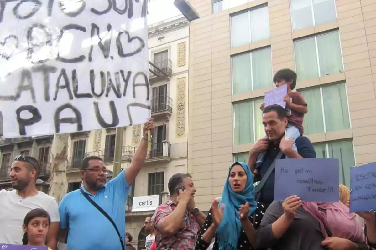 Varias personas participan en una manifestación en la calle, algunas sostienen pancartas y carteles con mensajes escritos a mano, mientras un niño está sentado sobre los hombros de un adulto.