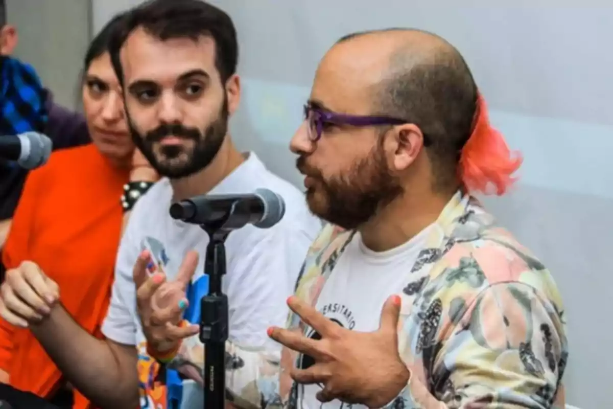 People participating in a panel discussion, one of them speaking into a microphone while the others listen alertly. People participating in a panel discussion, one of them speaking into a microphone while the others listen alertly.
