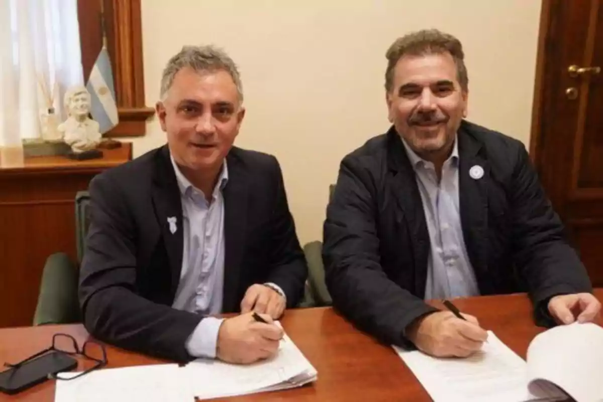 Dos hombres sentados en una oficina firmando documentos sobre una mesa, ambos sonríen y llevan traje, al fondo se ve una bandera de Argentina y un busto decorativo. Dos hombres sentados en una oficina firmando documentos sobre una mesa, ambos sonríen y llevan traje, al fondo se ve una bandera de Argentina y un busto decorativo.