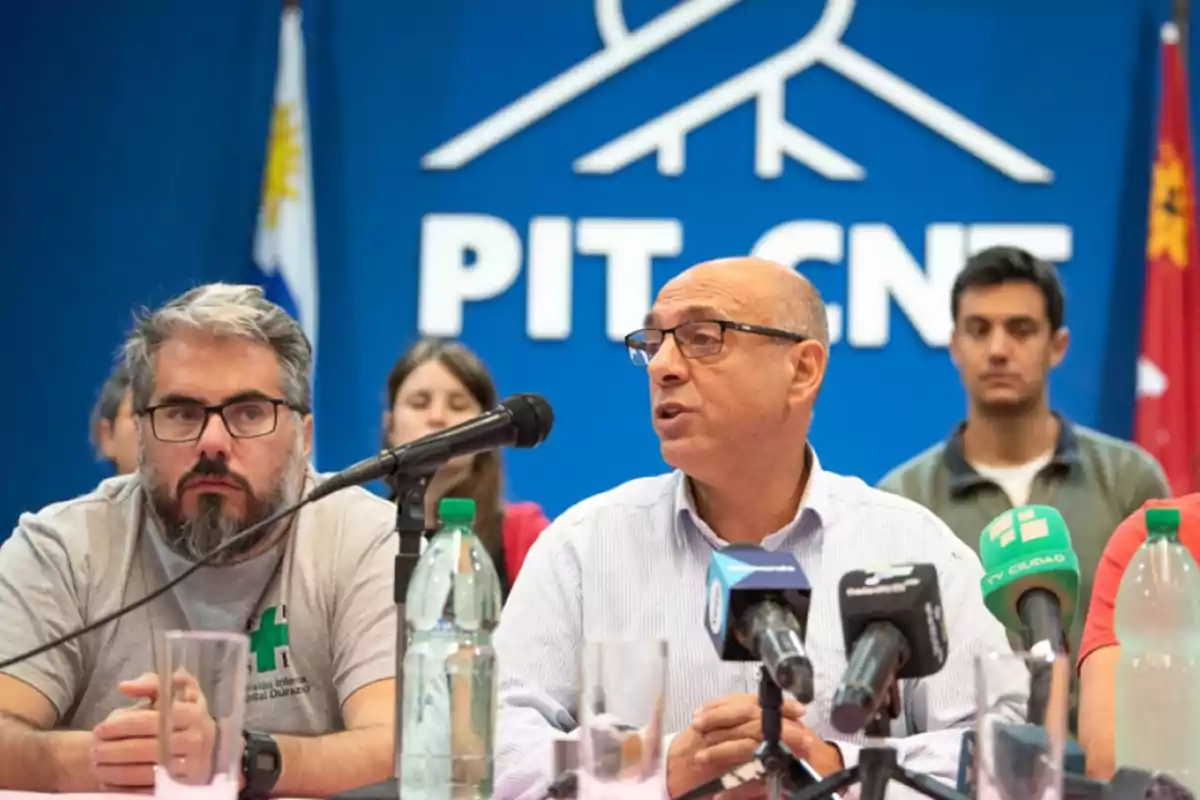 Hombres sentados en una mesa frente a micrófonos durante una conferencia de prensa con un fondo azul que tiene el logo de PIT-CNT y banderas a los costados Hombres sentados en una mesa frente a micrófonos durante una conferencia de prensa con un fondo azul que tiene el logo de PIT-CNT y banderas a los costados