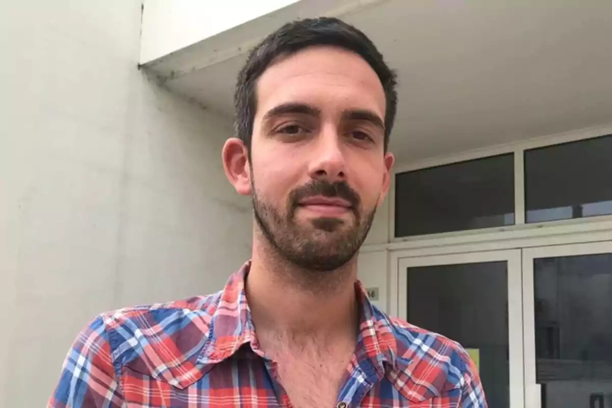 Hombre joven con barba y cabello corto oscuro vistiendo una camisa de cuadros rojos y azules frente a un edificio blanco