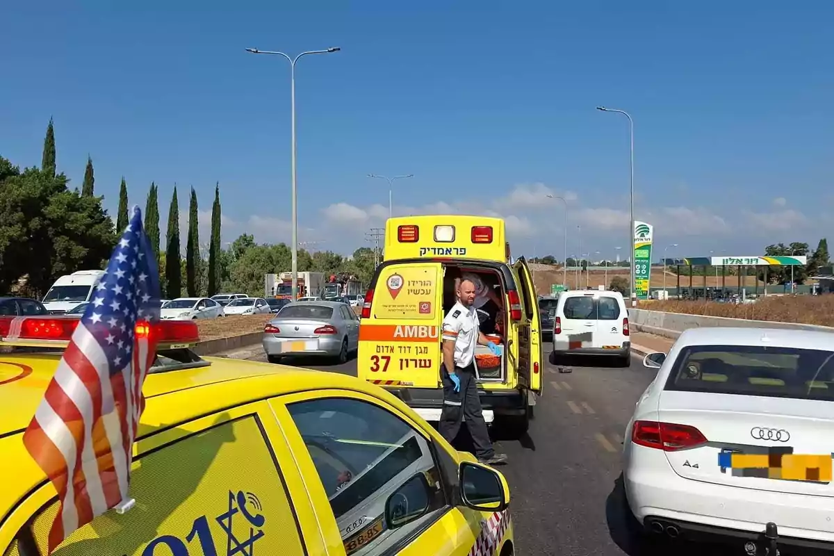Ambulancia amarilla detenida en una carretera junto a varios autos, un paramédico de pie junto a la puerta trasera abierta y una bandera de Estados Unidos en el vehículo de emergencia. Ambulancia amarilla detenida en una carretera junto a varios autos, un paramédico de pie junto a la puerta trasera abierta y una bandera de Estados Unidos en el vehículo de emergencia.
