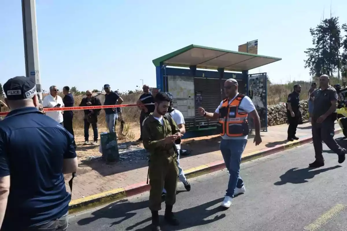 Several people, including police officers and a soldier, are at a cordoned-off scene near a bus stop.