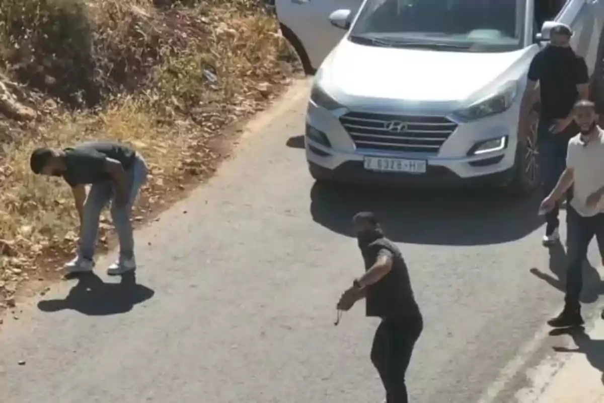 Un grupo de hombres en una carretera junto a un coche blanco, uno de ellos parece estar lanzando una piedra mientras otro recoge algo del suelo