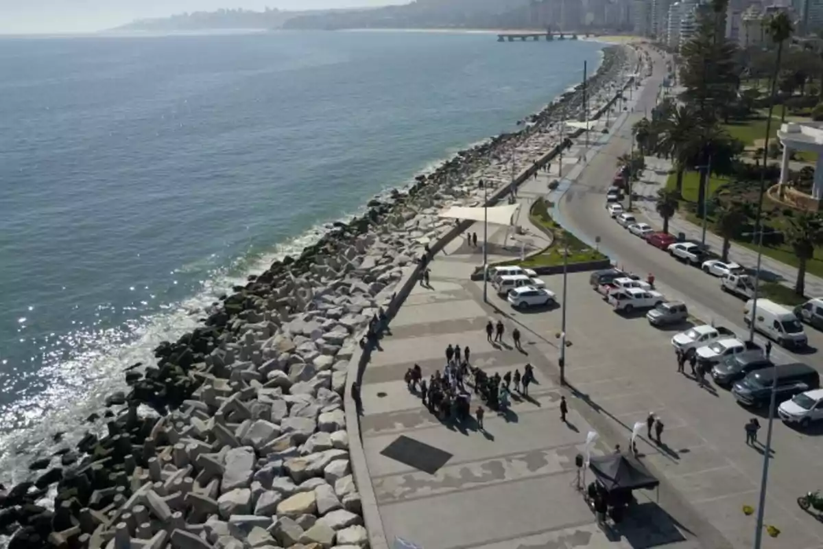 Vista aérea de un paseo costero junto al mar con un grupo de personas reunidas, autos estacionados y una ciudad al fondo