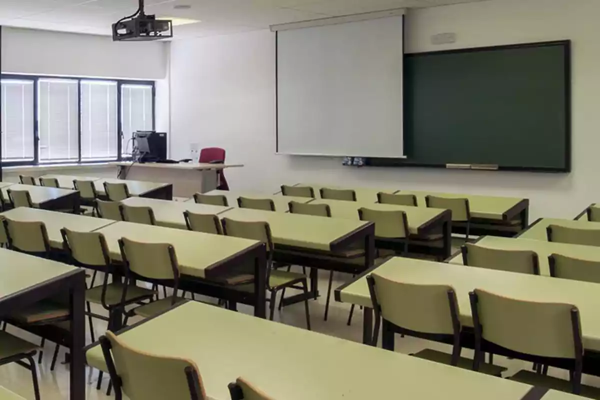 Aula vacía con escritorios y sillas alineados, un proyector en el techo, una pizarra y una pantalla de proyección junto al escritorio del profesor Aula vacía con escritorios y sillas alineados, un proyector en el techo, una pizarra y una pantalla de proyección junto al escritorio del profesor