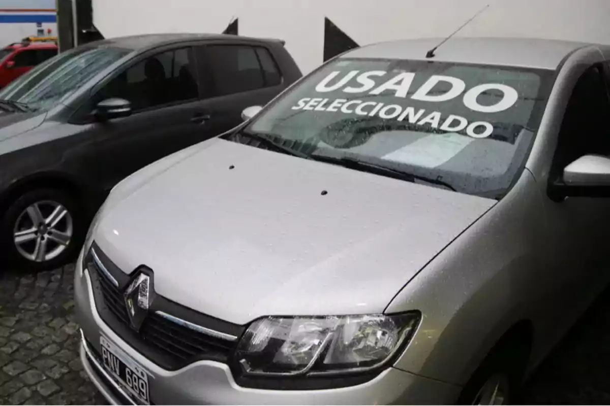 Auto Renault plateado con gotas de lluvia en el parabrisas y un cartel que dice usado seleccionado estacionado junto a otro vehículo en un lote de autos. Auto Renault plateado con gotas de lluvia en el parabrisas y un cartel que dice usado seleccionado estacionado junto a otro vehículo en un lote de autos.