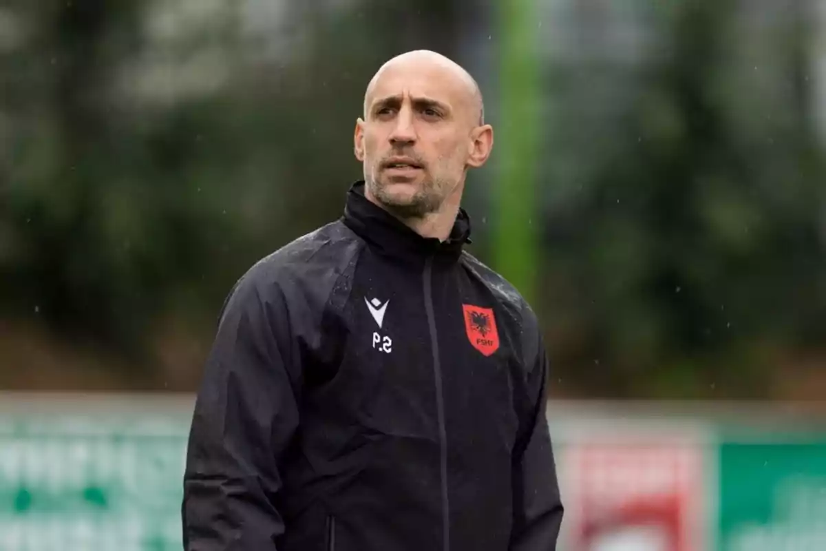 Bald man with a short beard wearing a black sports jacket with the Albanian Football Federation crest in an outdoor setting Bald man with a short beard wearing a black sports jacket with the Albanian Football Federation crest in an outdoor setting