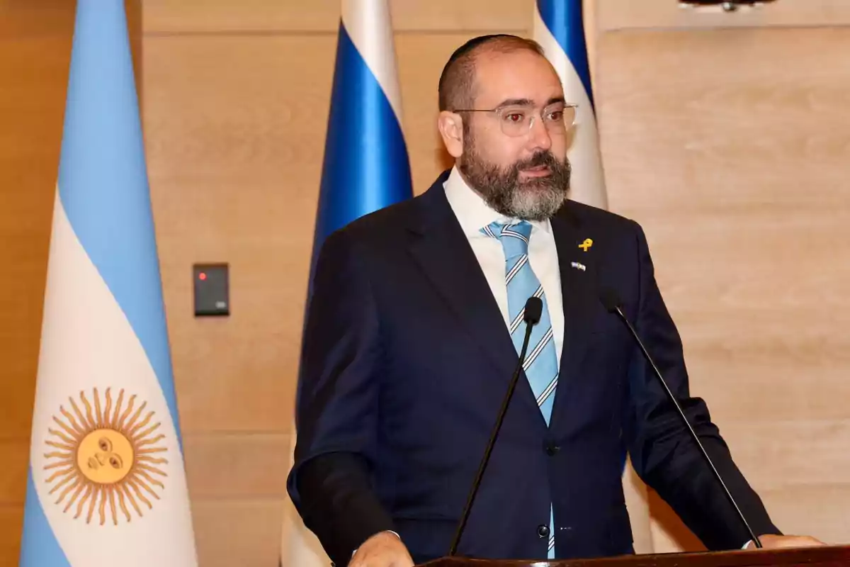 Hombre con barba y gafas hablando en un podio con micrófonos, vistiendo traje y corbata celeste, con banderas de Argentina e Israel de fondo