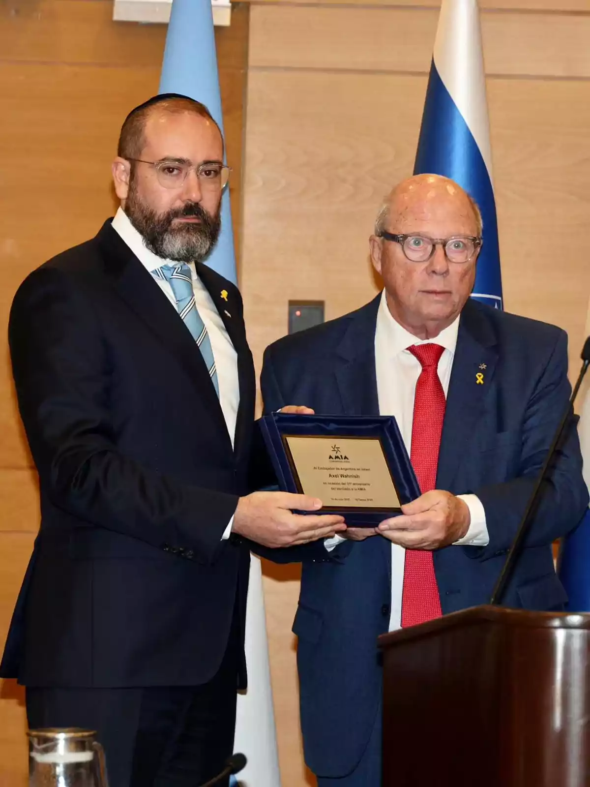 Dos hombres de traje posan juntos mientras sostienen una placa de reconocimiento en un evento formal con banderas de fondo Dos hombres de traje posan juntos mientras sostienen una placa de reconocimiento en un evento formal con banderas de fondo