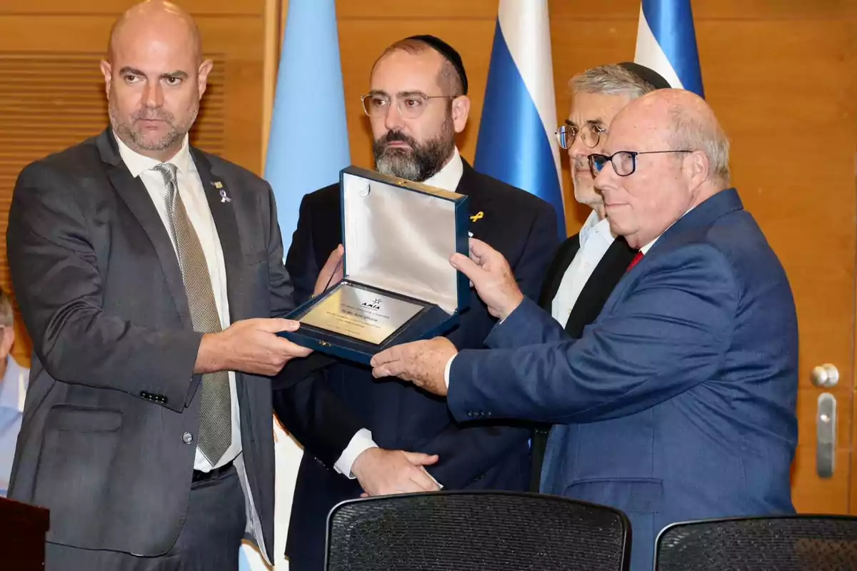 Cuatro hombres de traje sostienen una placa de reconocimiento en una ceremonia formal con banderas de fondo. Cuatro hombres de traje sostienen una placa de reconocimiento en una ceremonia formal con banderas de fondo.