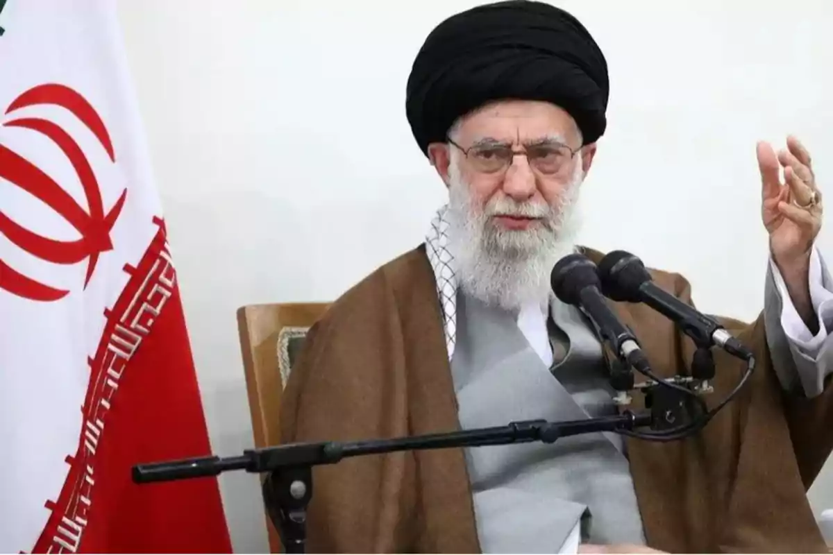 Hombre mayor con barba blanca y turbante negro hablando frente a micrófonos junto a una bandera de Irán Hombre mayor con barba blanca y turbante negro hablando frente a micrófonos junto a una bandera de Irán