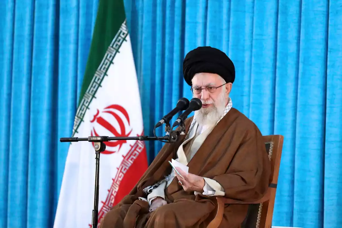 Elderly man with a beard and a black turban speaking in front of two microphones, seated next to an Iranian flag and a backdrop of blue curtains Elderly man with a beard and a black turban speaking in front of two microphones, seated next to an Iranian flag and a backdrop of blue curtains