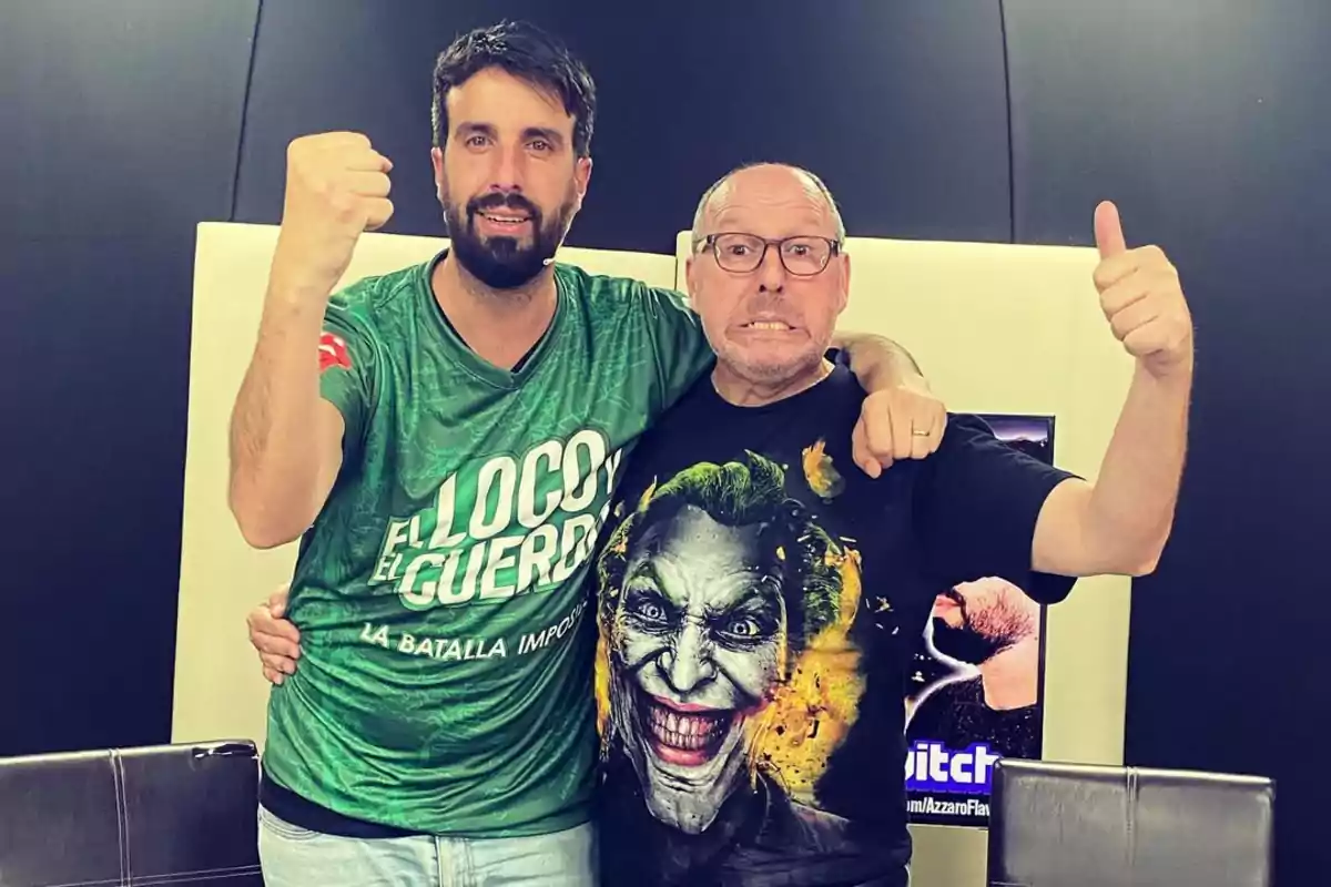 Dos hombres posan sonrientes y abrazados, uno lleva una camiseta verde con letras blancas y el otro una camiseta negra con la imagen del Joker, ambos levantan el puño en señal de celebración
