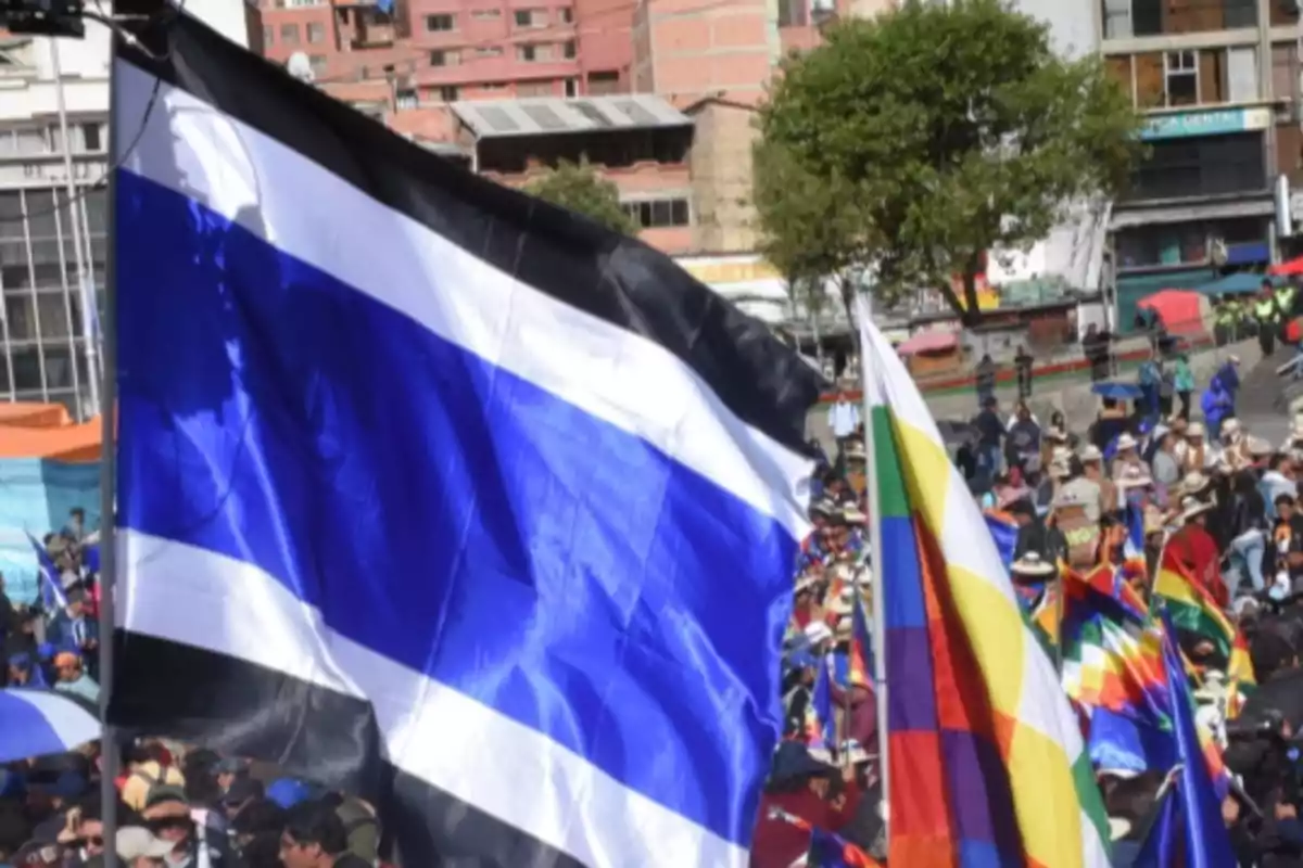 A blue, white, and black flag waves in the foreground while a crowd with other colorful flags gathers in an urban square. A blue, white, and black flag waves in the foreground while a crowd with other colorful flags gathers in an urban square.