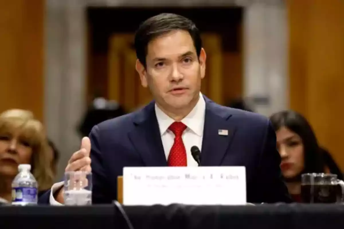 Dark-haired man in a blue suit and red tie speaking into a microphone during a conference or hearing with people in the background