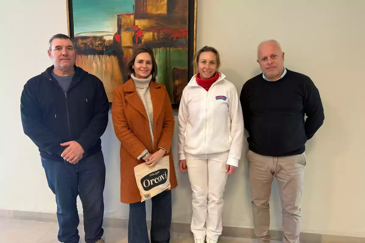 Cuatro personas posan de pie frente a una pared clara y un cuadro colorido, dos de ellas visten ropa casual y las otras dos llevan prendas más formales, una sostiene una bolsa con el logo Orcoví.