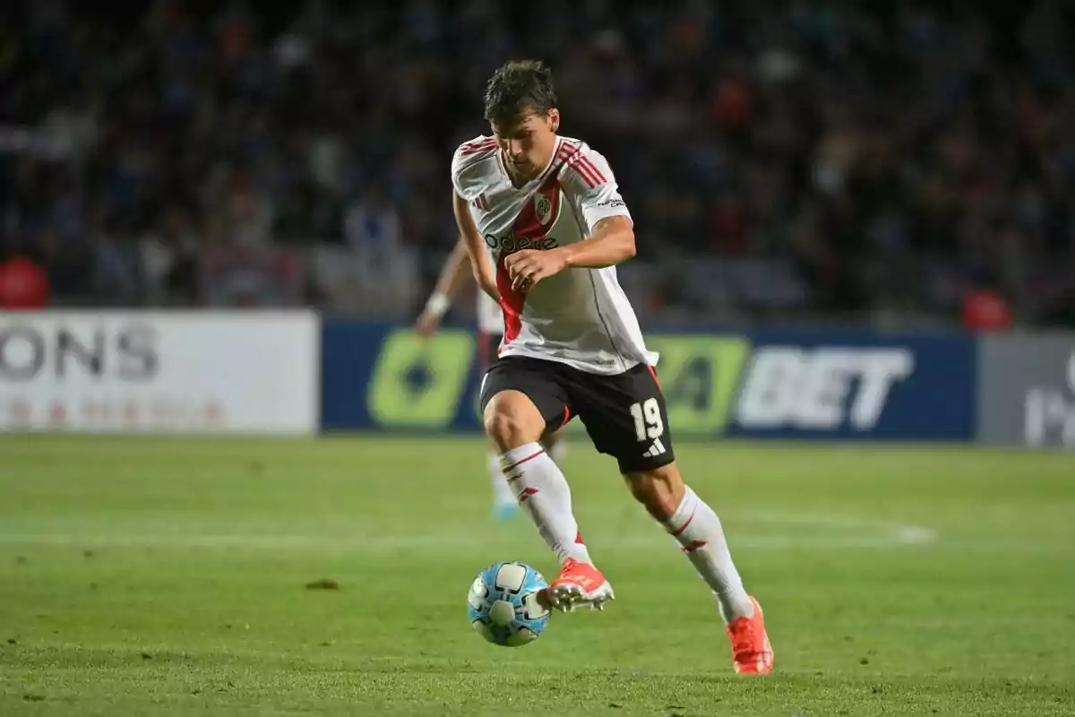 River Plate player wearing number 19 on his jersey controls the ball during a match in a lit-up stadium. River Plate player wearing number 19 on his jersey controls the ball during a match in a lit-up stadium.