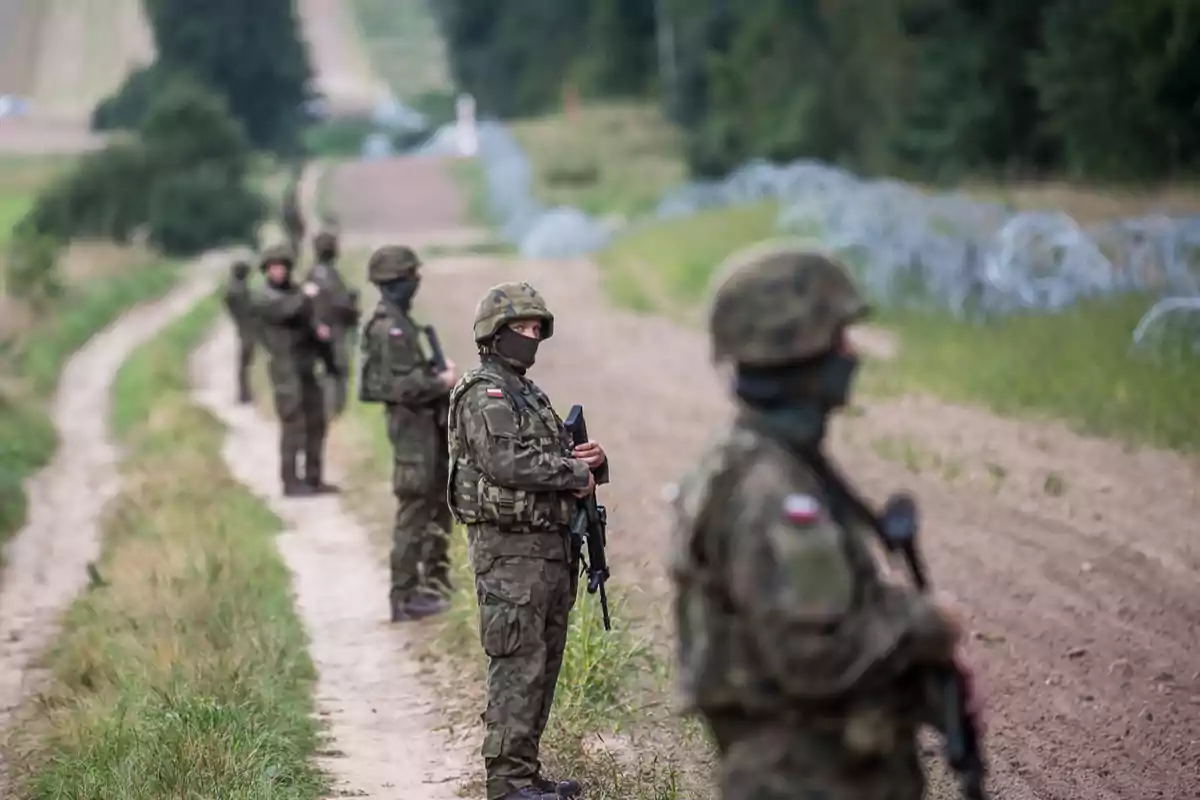 Soldiers in camouflage uniforms and armed with weapons patrol a rural area next to a wire fence on a dirt road. Soldiers in camouflage uniforms and armed with weapons patrol a rural area next to a wire fence on a dirt road.