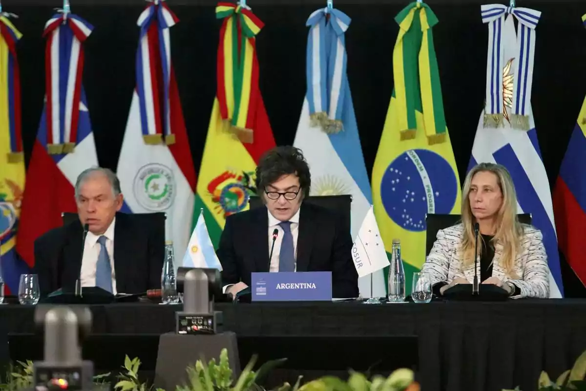 Tres personas sentadas en una mesa durante una conferencia internacional con banderas de varios países de fondo Tres personas sentadas en una mesa durante una conferencia internacional con banderas de varios países de fondo