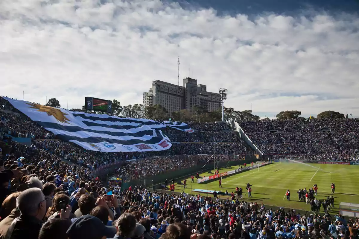 Stadium full of fans with a large Uruguay flag covering part of the stands during a soccer match on a sunny day