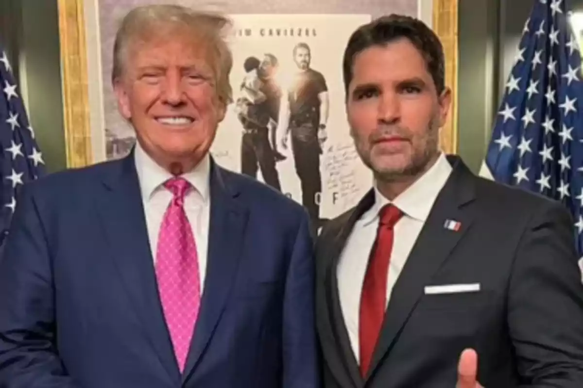 Two men in suits pose together in front of a poster and United States flags. Two men in suits pose together in front of a poster and United States flags.