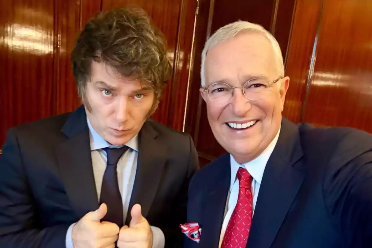 Two men dressed in suits pose together for a selfie; one of them smiles while the other gives a thumbs-up gesture, both standing in front of a wooden wall. Two men dressed in suits pose together for a selfie; one of them smiles while the other gives a thumbs-up gesture, both standing in front of a wooden wall.