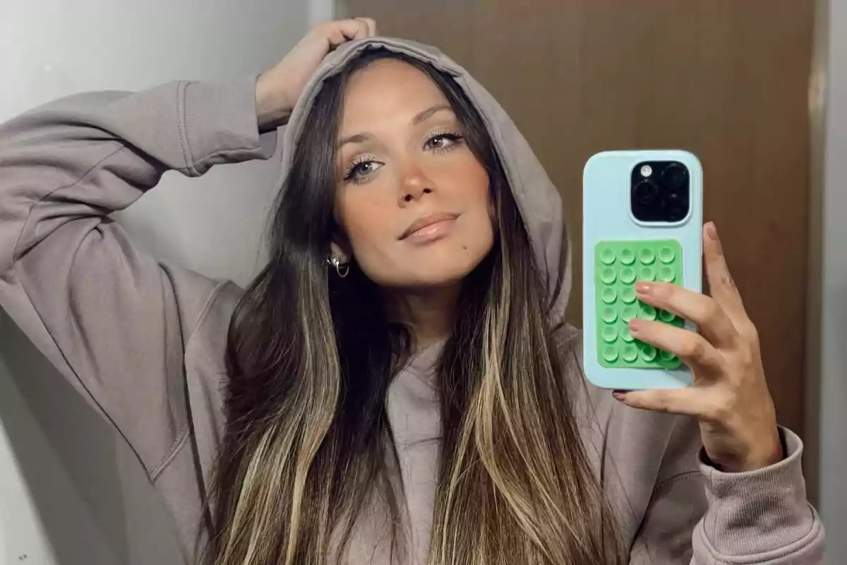 Mujer joven con sudadera beige y capucha tomándose una selfie frente al espejo con un teléfono de funda azul y verde