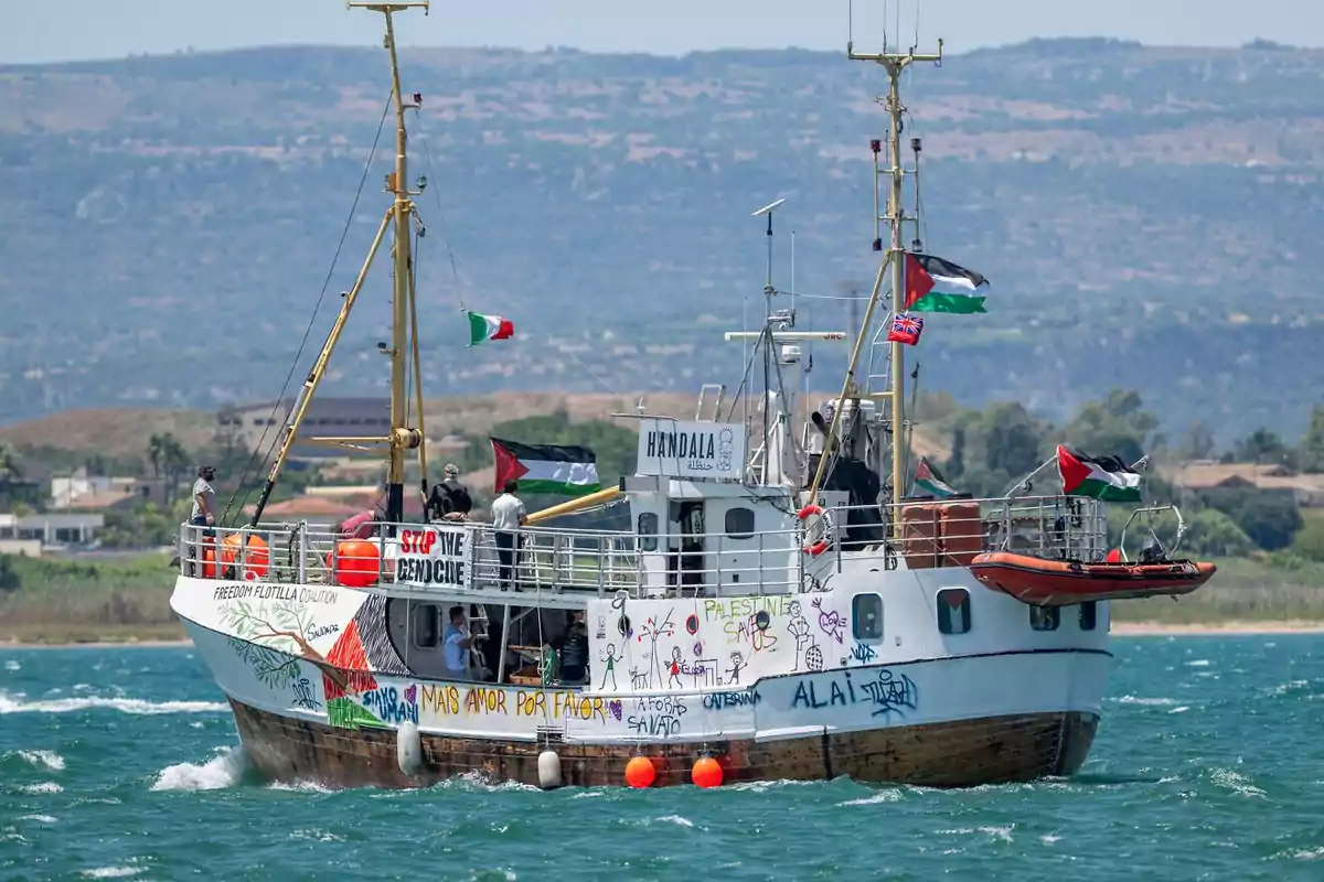 Barco con banderas palestinas y mensajes pintados navegando cerca de la costa con varias personas a bordo