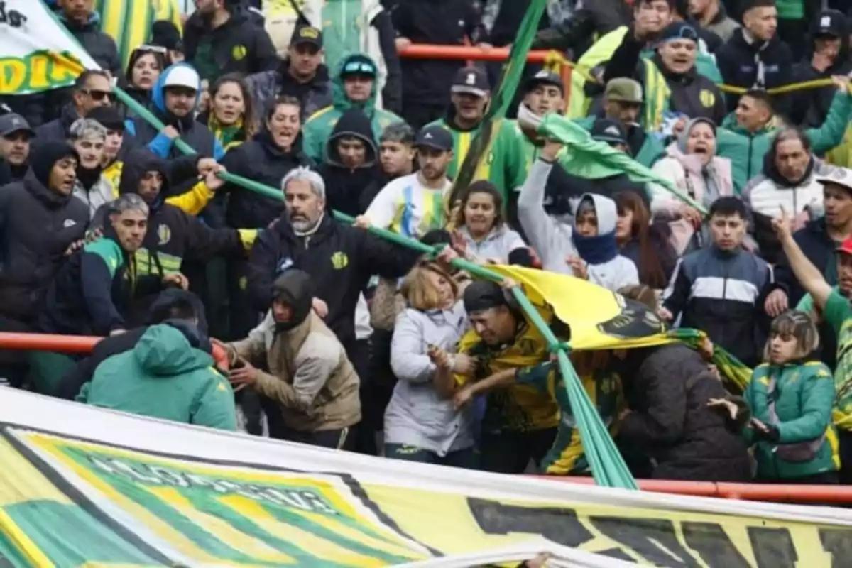 Aficionados de fútbol discuten y se empujan en las gradas durante un partido, algunos sostienen banderas y otros intentan separarlos mientras el ambiente se muestra tenso.
