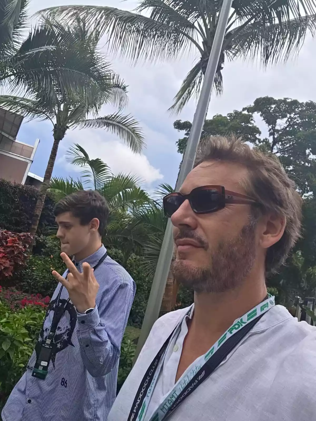Two people are walking outdoors among palm trees and vegetation, both wearing badges around their necks, and one of them is wearing sunglasses. Two people are walking outdoors among palm trees and vegetation, both wearing badges around their necks, and one of them is wearing sunglasses.