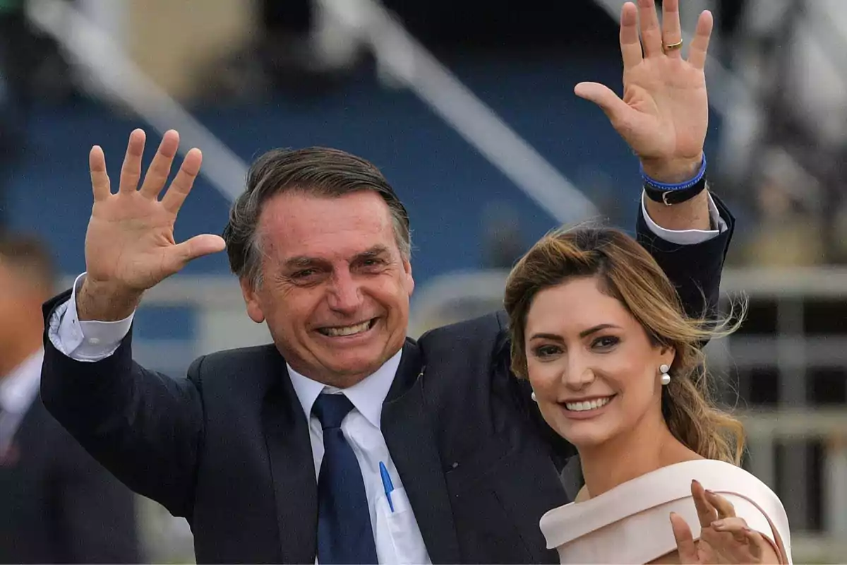 Un hombre y una mujer sonríen y saludan levantando las manos en un evento público