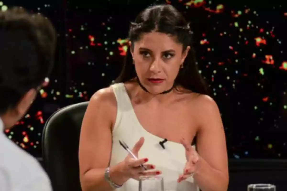 Long-haired, dark-haired woman in a one-shoulder white dress speaking and gesturing during an interview in a studio with a dark background and colored lights Long-haired, dark-haired woman in a one-shoulder white dress speaking and gesturing during an interview in a studio with a dark background and colored lights