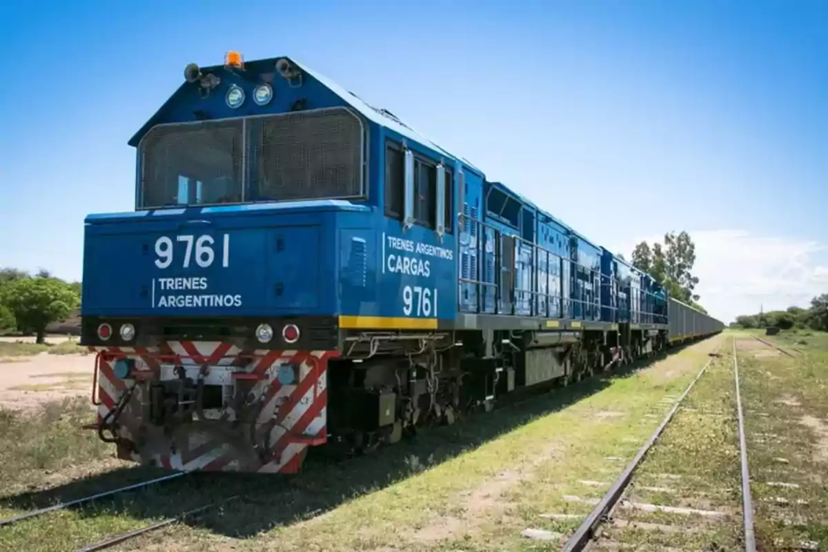 Tren de carga azul de Trenes Argentinos circulando por una vía en un entorno rural bajo un cielo despejado