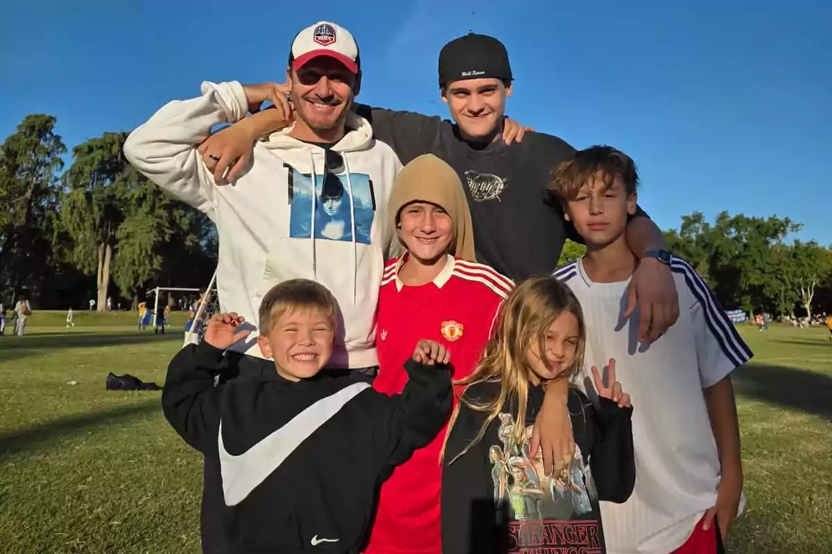 Grupo de cinco niños y un adulto posando sonrientes en un campo de césped bajo un cielo despejado Grupo de cinco niños y un adulto posando sonrientes en un campo de césped bajo un cielo despejado