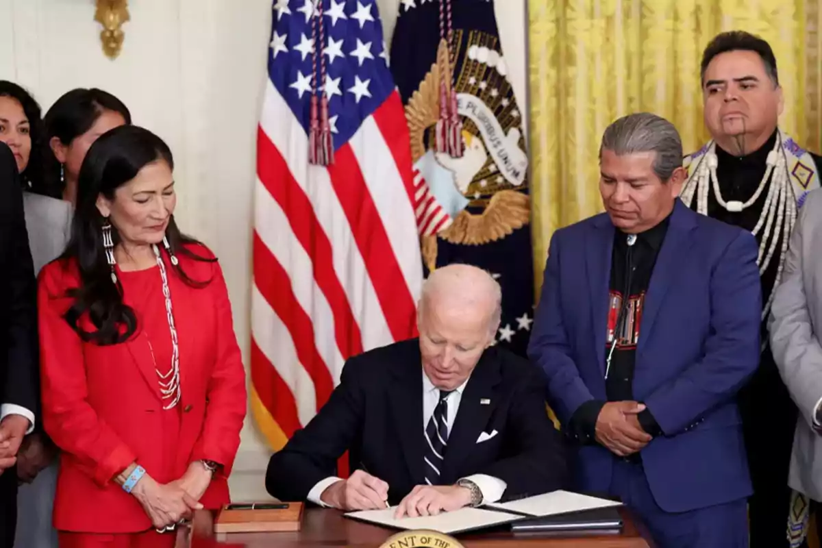 Un hombre de cabello canoso firma un documento en una mesa mientras varias personas lo rodean y observan, con banderas estadounidenses y un escudo presidencial al fondo