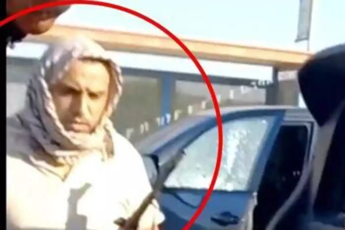 Hombre con la cabeza cubierta sostiene un objeto frente a un automóvil con la ventana rota, rodeado por un círculo rojo