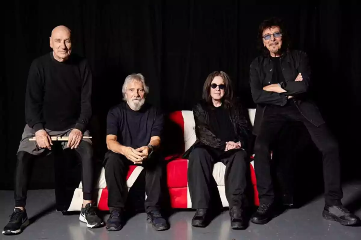 Four men dressed in black pose seated on a couch with a dark background. Four men dressed in black pose seated on a couch with a dark background.