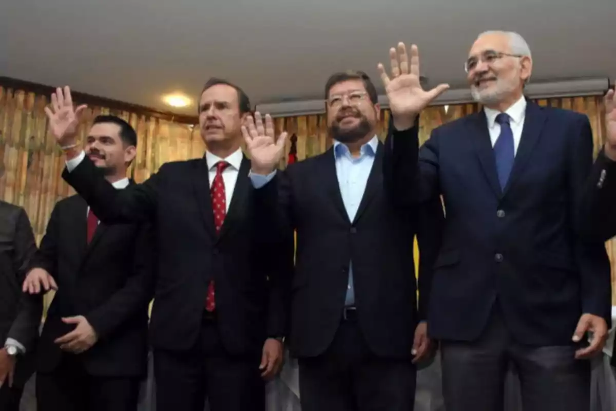 Cinco hombres vestidos con traje y corbata levantan la mano y sonríen en un ambiente interior con cortinas de fondo