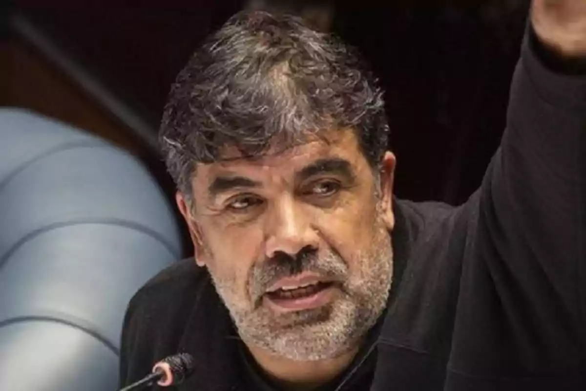 Hombre de cabello y barba canosos hablando frente a un micrófono con el brazo levantado