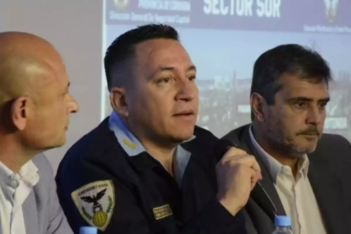 Three men sitting at a table during a conference, one of them wearing a police uniform and speaking while the other two listen alertly.