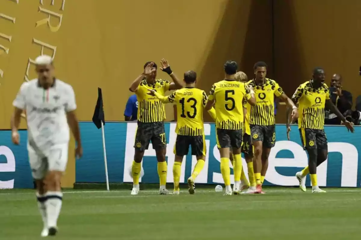 Jugadores de un equipo de fútbol con uniforme amarillo y negro celebran un gol cerca de la esquina del campo mientras un jugador rival camina cabizbajo en primer plano. Jugadores de un equipo de fútbol con uniforme amarillo y negro celebran un gol cerca de la esquina del campo mientras un jugador rival camina cabizbajo en primer plano.