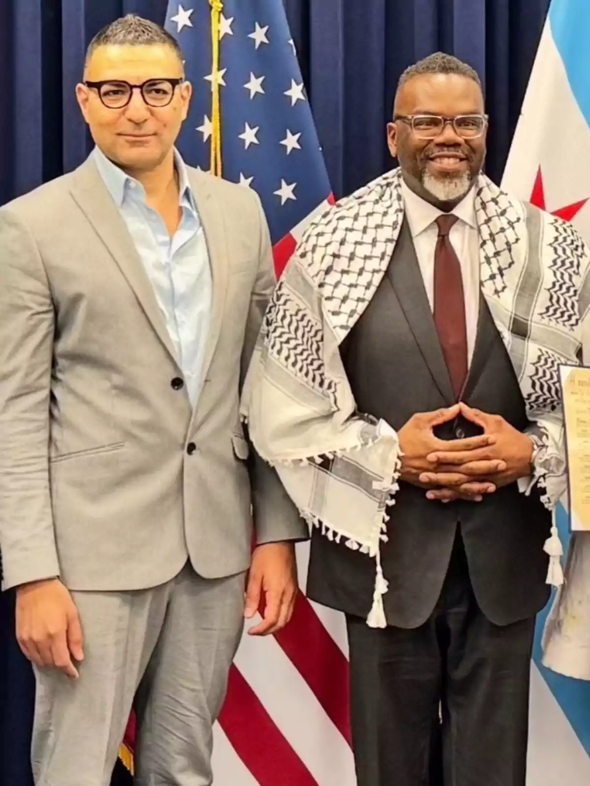 Dos hombres posan juntos frente a banderas de Estados Unidos y Chicago uno de ellos lleva un traje gris claro y gafas mientras que el otro viste un traje oscuro con corbata y una kufiya sobre los hombros ambos sonríen a la cámara