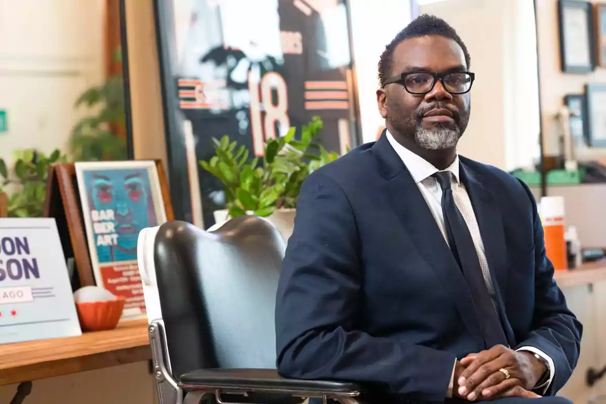 Man with glasses and a graying beard wearing a navy blue suit sitting in a barbershop with plants and pictures in the background