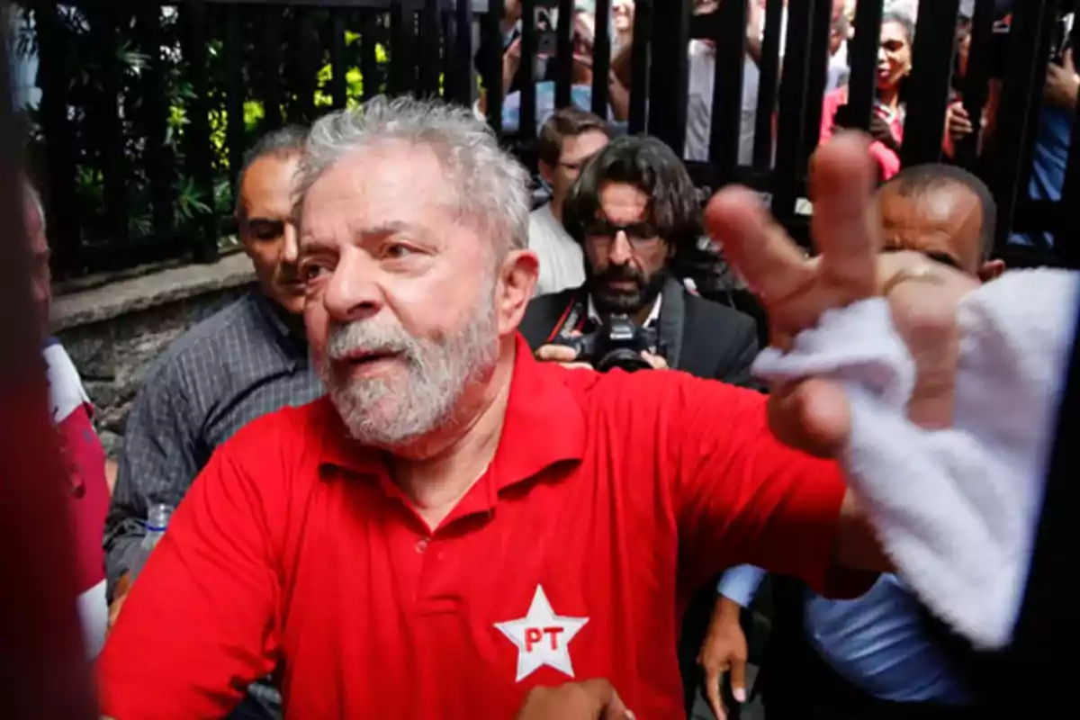 Gray-bearded man wearing a red jersey with a PT logo, surrounded by people and photographers in an outdoor setting Gray-bearded man wearing a red jersey with a PT logo, surrounded by people and photographers in an outdoor setting